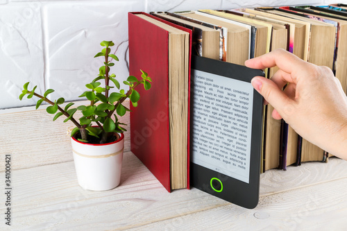 Woman hand takes e-reader from shelf with paper books and small potted plant. Copy space on e-book display. E-reading for pleasure and education. photo