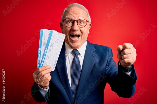 Senior grey haired business man holding airplane boarding pass over red background screaming proud and celebrating victory and success very excited, cheering emotion