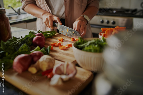 Fototapeta Naklejka Na Ścianę i Meble -  Closeup of young female hands chopping fresh vegetables on chopping board while in modern kitchen - preparing a healthy meal to boost immune system and fight off coronavirus