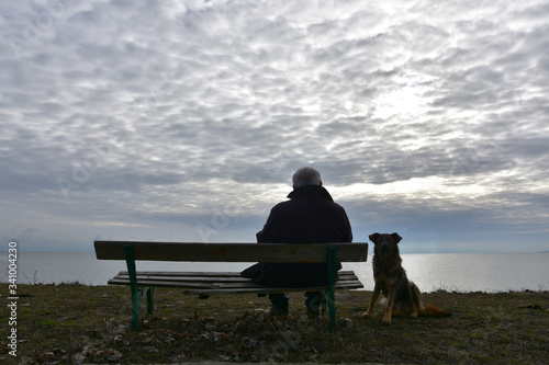 Lonely old man with dog. Senior man sitting on an old wooden bench and watching dark winter see