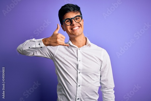 Young handsome business man wearing shirt and glasses over isolated purple background smiling doing phone gesture with hand and fingers like talking on the telephone. Communicating concepts.