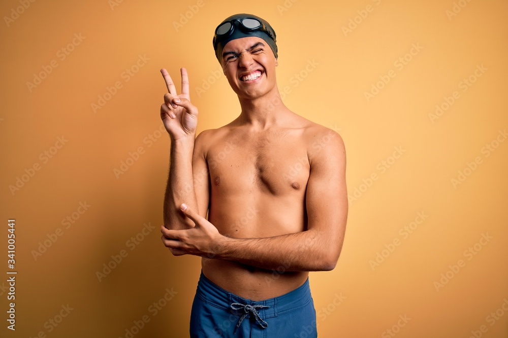 Young handsome man shirtless wearing swimsuit and swim cap over isolated yellow background smiling with happy face winking at the camera doing victory sign with fingers. Number two.
