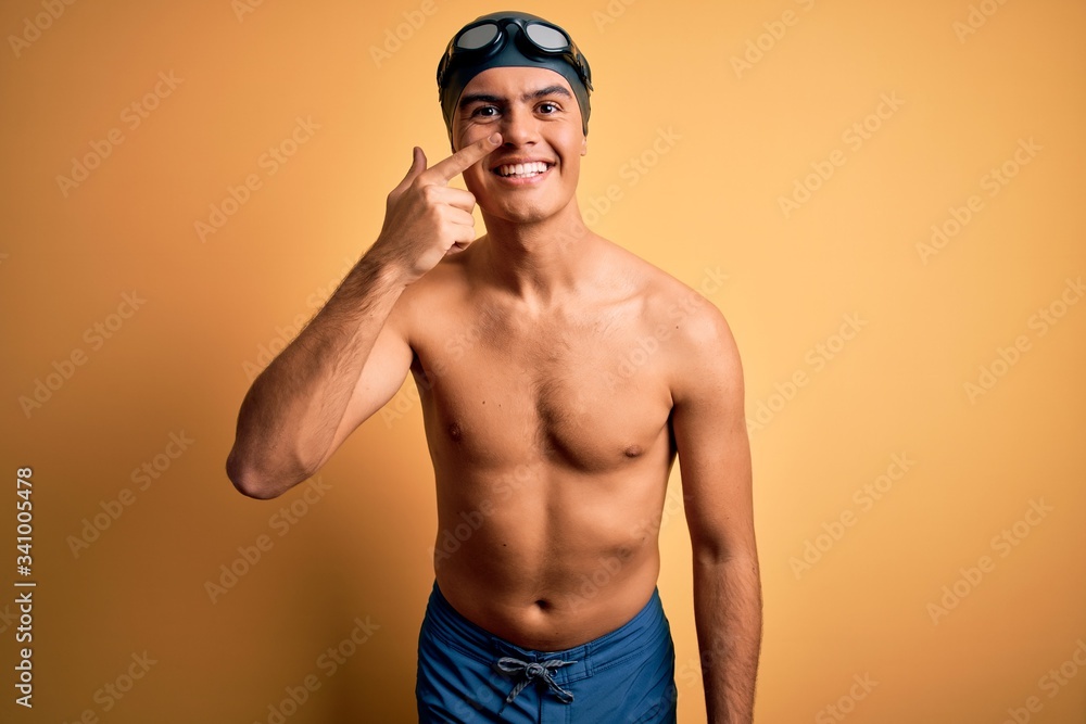 Young handsome man shirtless wearing swimsuit and swim cap over isolated yellow background Pointing with hand finger to face and nose, smiling cheerful. Beauty concept