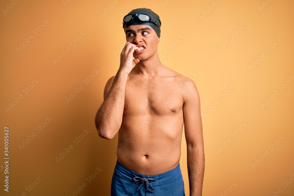 Young handsome man shirtless wearing swimsuit and swim cap over isolated yellow background looking stressed and nervous with hands on mouth biting nails. Anxiety problem.