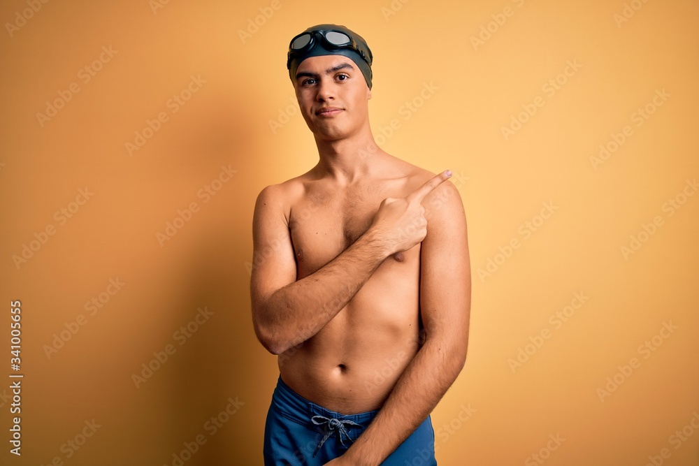 Young handsome man shirtless wearing swimsuit and swim cap over isolated yellow background Pointing with hand finger to the side showing advertisement, serious and calm face
