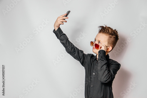 Tableau sur toile a fashionable guy in sunglasses and a black shirt with a cool hairstyle looks at the phone and photographs himself on a white background