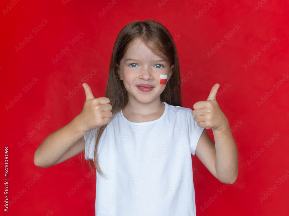 Little girl in a white T-shirt with the Polish flag on her cheek shows thumb up gesture, concept of learning the Polish language, immigration to Poland