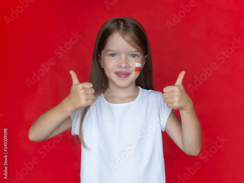 Obraz na płótnie Little girl in a white T-shirt with the Polish flag on her cheek shows thumb up gesture, concept of learning the Polish language, immigration to Poland w salonie