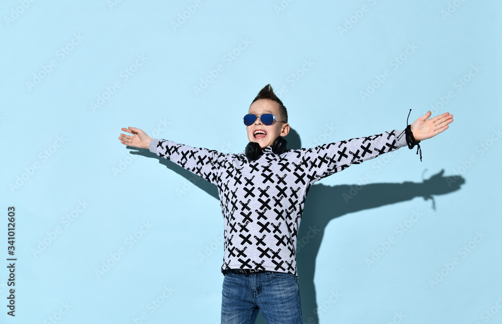 Teenager with headphones on neck, in sunglasses, blue jeans and stylish hoodie. Spreaded hands, smiling, posing on blue background