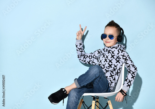 Teenage kid in headphones, sunglasses, blue jeans, hoodie, sneakers. Showing victory sign, sitting on white chair. Blue background photo