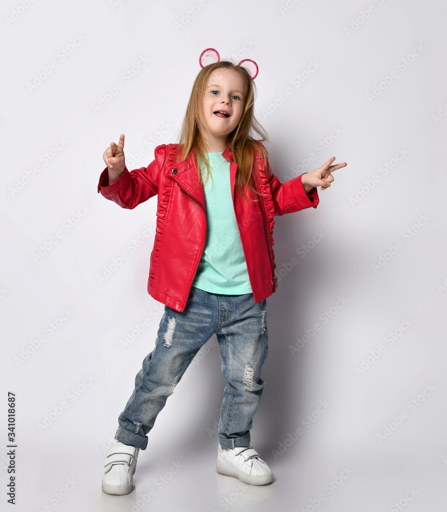Cool and positive blond baby girl kid in red leather jacket, white t-shirt and blue jeans gestures V-signs victory Cool and positive blond baby girl kid in red leather jacket, white t-shirt and blue jeans gestures V-signs victory