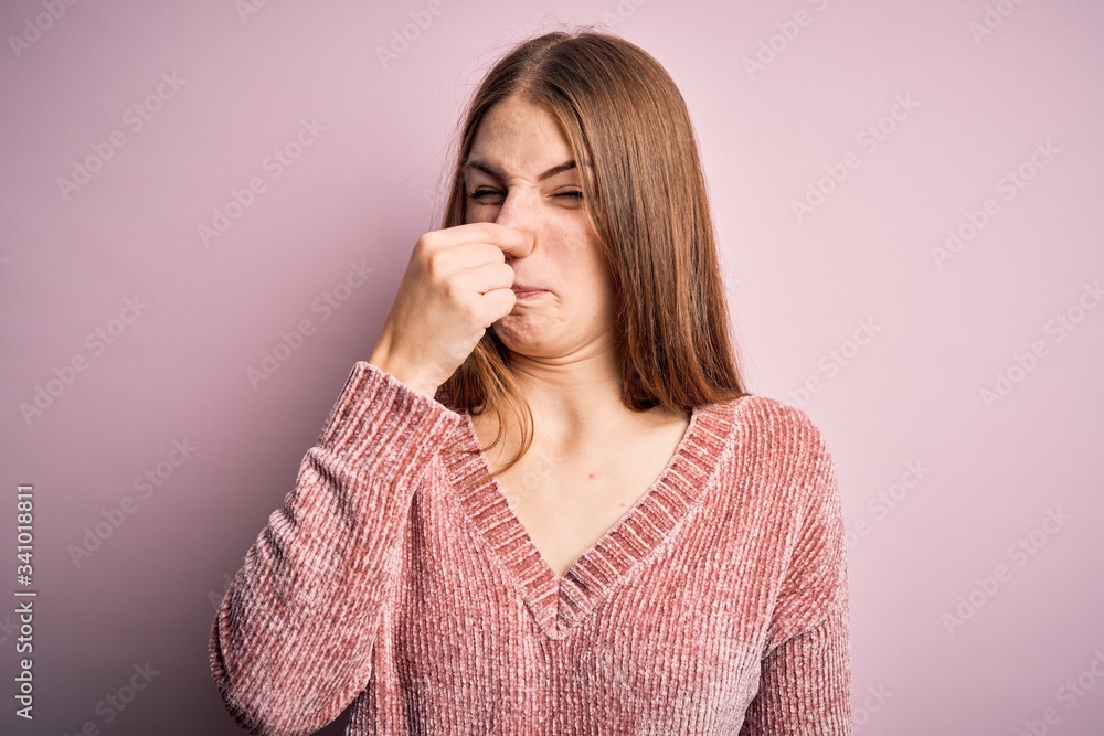 Young beautiful redhead woman wearing casual sweater over isolated pink background smelling something stinky and disgusting, intolerable smell, holding breath with fingers on nose. Bad smell Young beautiful redhead woman wearing casual sweater over isolated pink background smelling something stinky and disgusting, intolerable smell, holding breath with fingers on nose. Bad smell