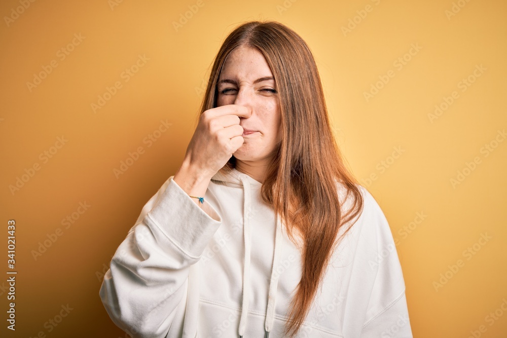 Young beautiful redhead sporty woman wearing sweatshirt over isolated yellow background smelling something stinky and disgusting, intolerable smell, holding breath with fingers on nose. Bad smell Young beautiful redhead sporty woman wearing sweatshirt over isolated yellow background smelling something stinky and disgusting, intolerable smell, holding breath with fingers on nose. Bad smell