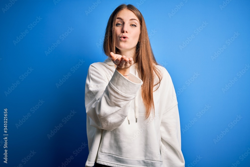 Young beautiful redhead sporty woman wearing sweatshirt over isolated blue background looking at the camera blowing a kiss with hand on air being lovely and sexy. Love expression. Young beautiful redhead sporty woman wearing sweatshirt over isolated blue background looking at the camera blowing a kiss with hand on air being lovely and sexy. Love expression.