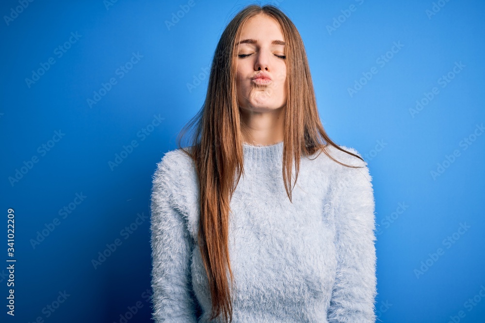 Young beautiful redhead woman wearing casual sweater over isolated blue background looking at the camera blowing a kiss on air being lovely and sexy. Love expression. Young beautiful redhead woman wearing casual sweater over isolated blue background looking at the camera blowing a kiss on air being lovely and sexy. Love expression.