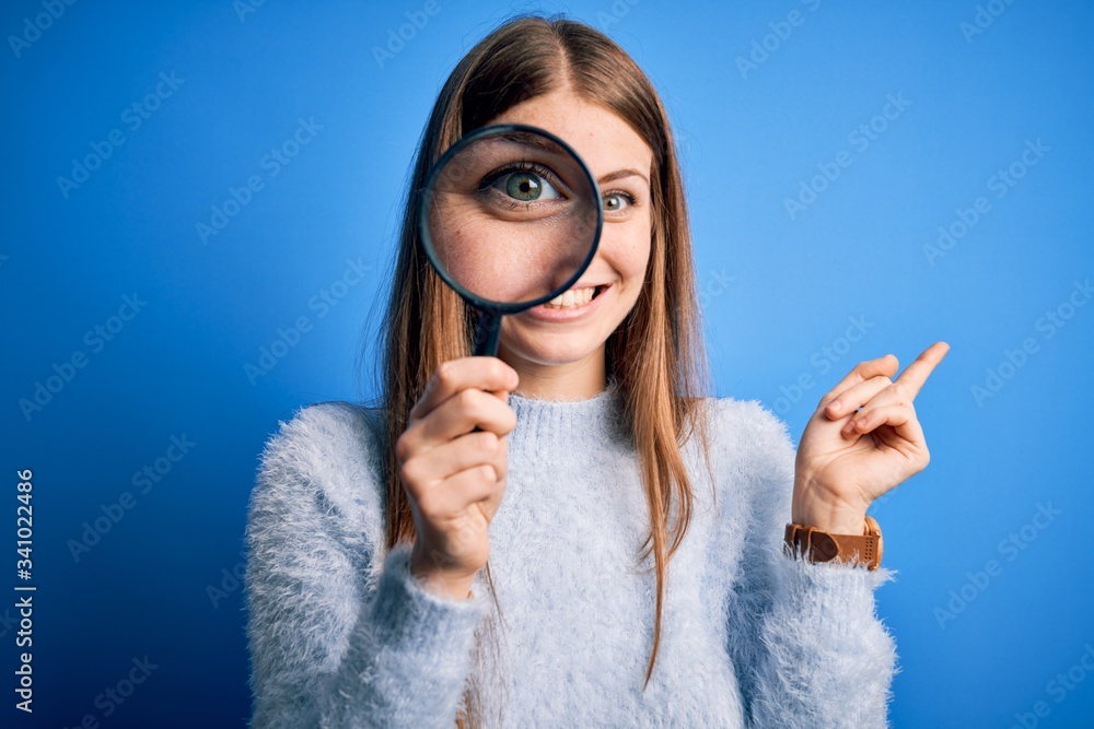 Young beautiful redhead detective woman using magnifying glass over isolated blue background very happy pointing with hand and finger to the side Young beautiful redhead detective woman using magnifying glass over isolated blue background very happy pointing with hand and finger to the side