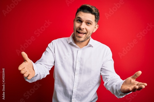 Young business man with blue eyes wearing elegant shirt standing over red isolated background smiling cheerful offering hands giving assistance and acceptance. photo