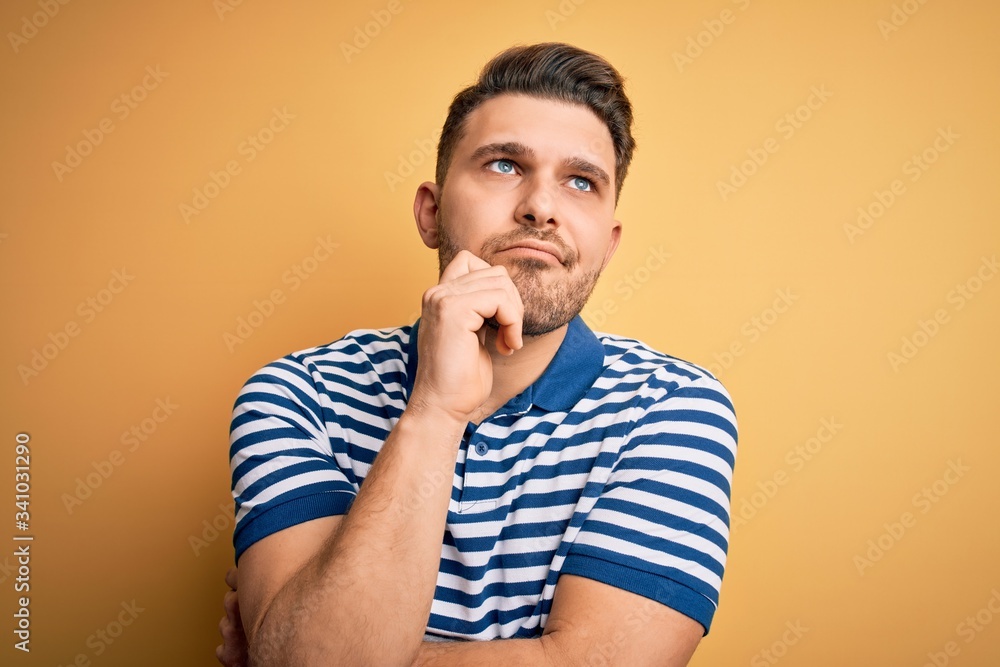 Young man with blue eyes wearing casual striped t-shirt over yellow background with hand on chin thinking about question, pensive expression. Smiling with thoughtful face. Doubt concept. Young man with blue eyes wearing casual striped t-shirt over yellow background with hand on chin thinking about question, pensive expression. Smiling with thoughtful face. Doubt concept.