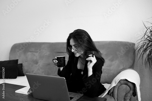 Fashionable young Caucasian woman sitting on sofa at home and working from home, remotely. She is keyboarding on laptop, drinking coffee and taking notes. Freelance work concept. black and white photo