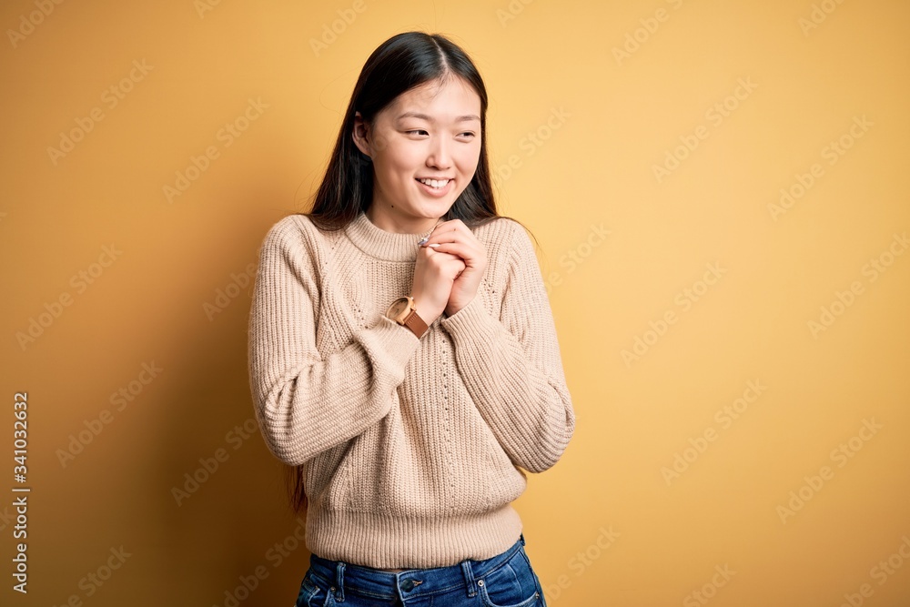 Young beautiful asian woman wearing casual sweater over yellow isolated background laughing nervous and excited with hands on chin looking to the side Young beautiful asian woman wearing casual sweater over yellow isolated background laughing nervous and excited with hands on chin looking to the side