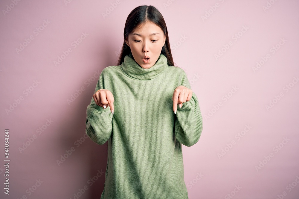 Young beautiful asian woman wearing green winter sweater over pink solated background Pointing down with fingers showing advertisement, surprised face and open mouth Young beautiful asian woman wearing green winter sweater over pink solated background Pointing down with fingers showing advertisement, surprised face and open mouth