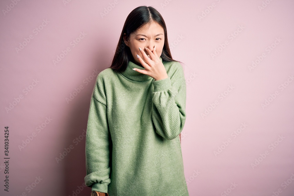 Young beautiful asian woman wearing green winter sweater over pink solated background smelling something stinky and disgusting, intolerable smell, holding breath with fingers on nose. Bad smell Young beautiful asian woman wearing green winter sweater over pink solated background smelling something stinky and disgusting, intolerable smell, holding breath with fingers on nose. Bad smell