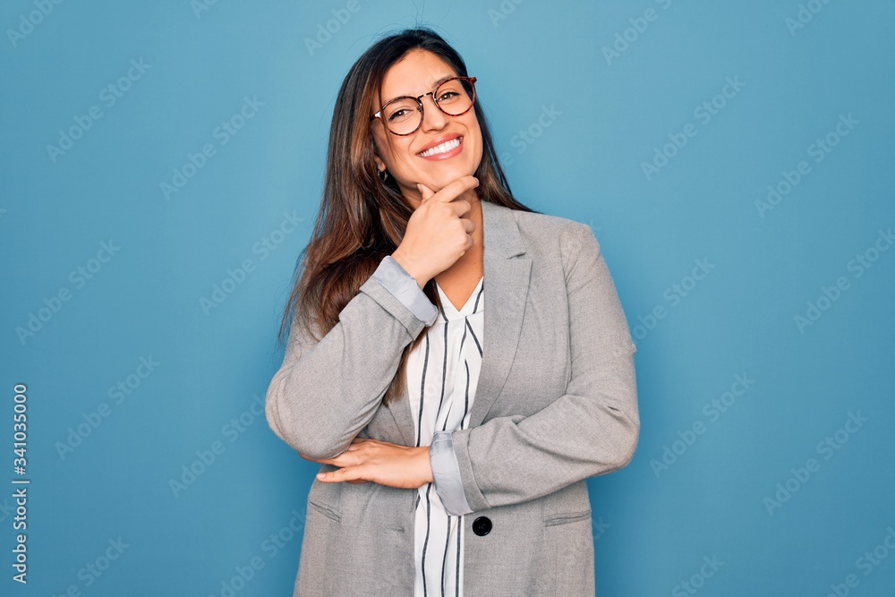 Young hispanic business woman wearing glasses standing over blue isolated background looking confident at the camera smiling with crossed arms and hand raised on chin. Thinking positive. Young hispanic business woman wearing glasses standing over blue isolated background looking confident at the camera smiling with crossed arms and hand raised on chin. Thinking positive.