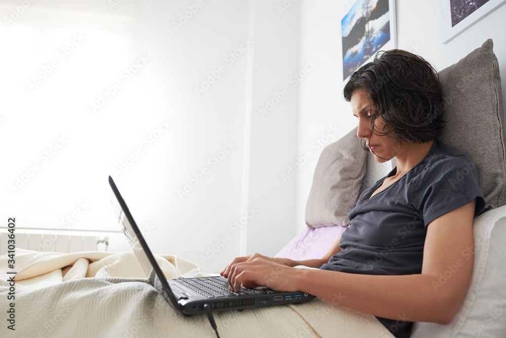 
Woman in bed with a laptop and books working from home. She is on a minimalist white floor.