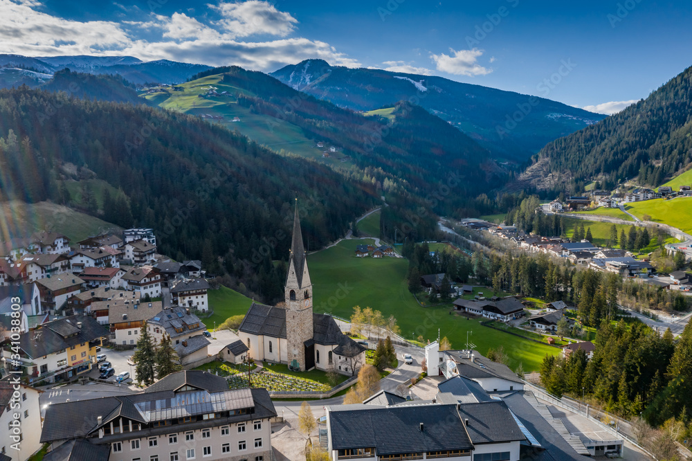 Aerial view of valley and small city with church, green slopes of the mountains of Italy, Trentino, San Martino in Badia, roofs of houses of settlements, green meadows, Dolomites on background,