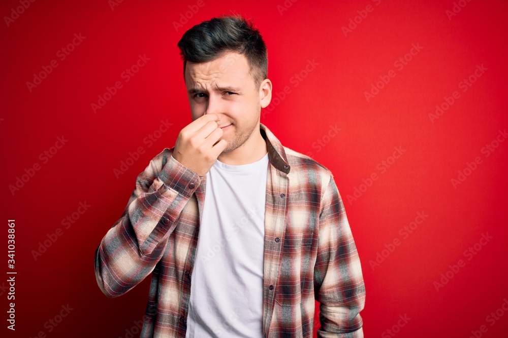 Young handsome caucasian man wearing casual modern shirt over red isolated background smelling something stinky and disgusting, intolerable smell, holding breath with fingers on nose. Bad smell