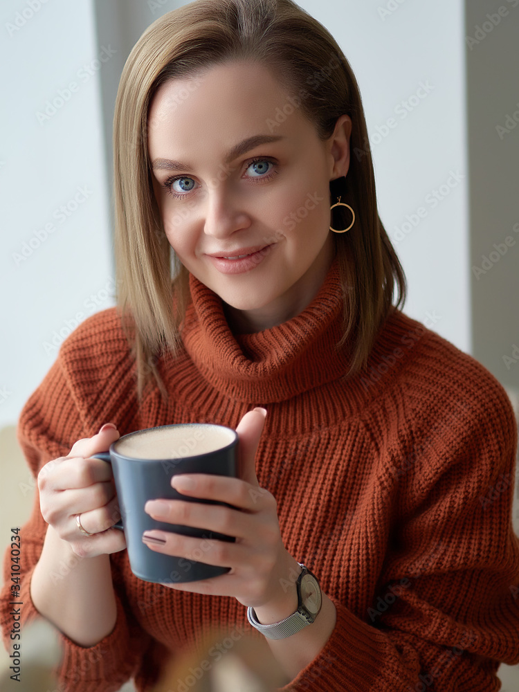 a positive girl in a brown sweater sits at the window and drinks coffee. a smile on his face. a positive girl in a brown sweater sits at the window and drinks coffee. a smile on his face.