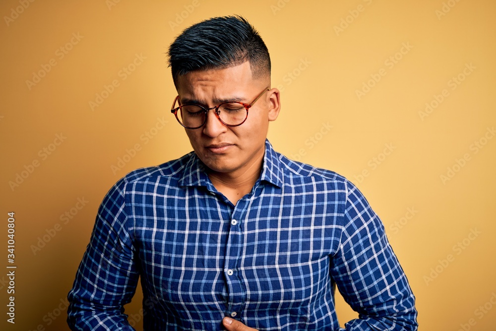 Young handsome latin man wearing casual shirt and glasses over yellow background with hand on stomach because indigestion, painful illness feeling unwell. Ache concept. Young handsome latin man wearing casual shirt and glasses over yellow background with hand on stomach because indigestion, painful illness feeling unwell. Ache concept.