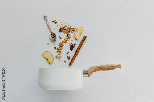 Apples, oat granola, cinnamon stick, pumpkin seeds, dried berries and syrup going into a white saucepan on a pastel white background