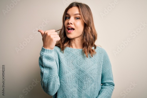 Young blonde girl wearing casual blue winter sweater over isolated background smiling with happy face looking and pointing to the side with thumb up.