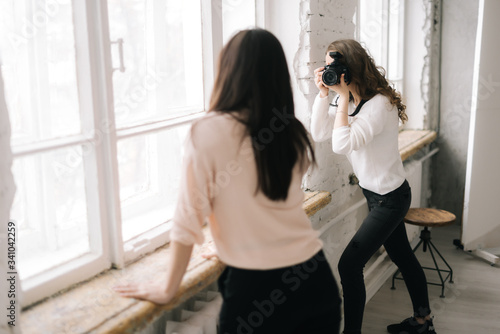 Young professional woman model posing in photo studio for female photographer near a window while she is shooting with digital camera. Concept of creative work in photo studio, backstage job.
