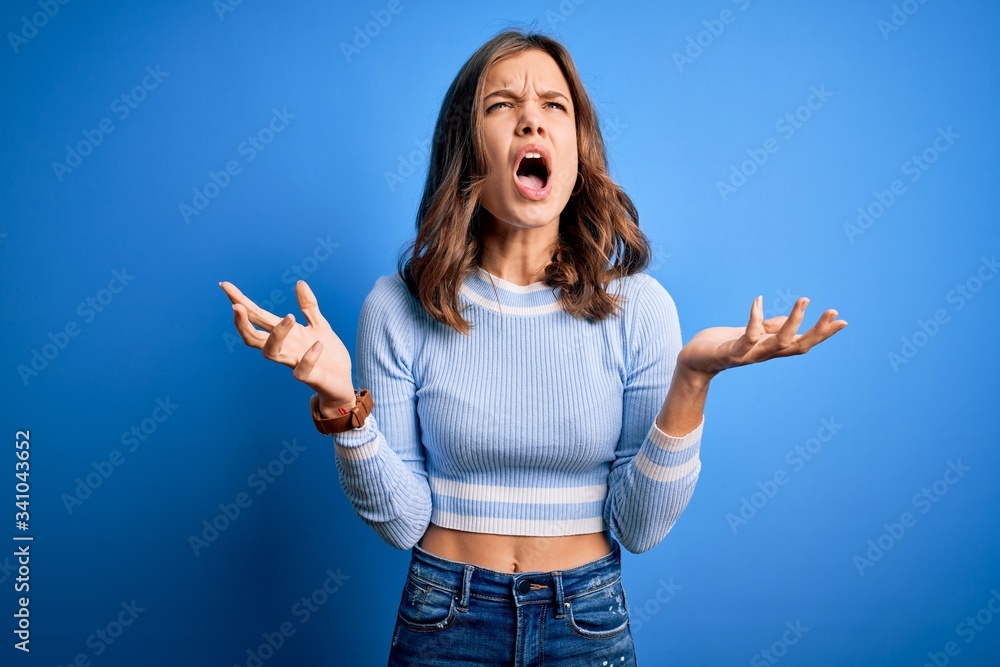 Young beautiful blonde girl wearing casual sweater standing over blue isolated background crazy and mad shouting and yelling with aggressive expression and arms raised. Frustration concept. Young beautiful blonde girl wearing casual sweater standing over blue isolated background crazy and mad shouting and yelling with aggressive expression and arms raised. Frustration concept.