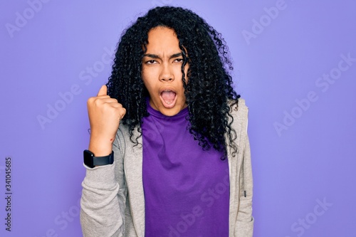 Young african american sporty woman wearing casual sweatshirt over purple background angry and mad raising fist frustrated and furious while shouting with anger. Rage and aggressive concept. © Krakenimages.com