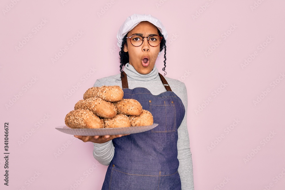 Young african american baker woman wearing apron holding wholemeal bread with cereal scared in shock with a surprise face, afraid and excited with fear expression Young african american baker woman wearing apron holding wholemeal bread with cereal scared in shock with a surprise face, afraid and excited with fear expression