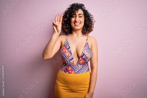 Young beautiful arab woman on vacation wearing swimsuit and sunglasses over pink background showing and pointing up with fingers number three while smiling confident and happy.