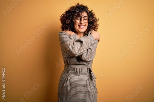 Canvastavla Beautiful arab business woman wearing dress and glasses standing over yellow background Hugging oneself happy and positive, smiling confident
