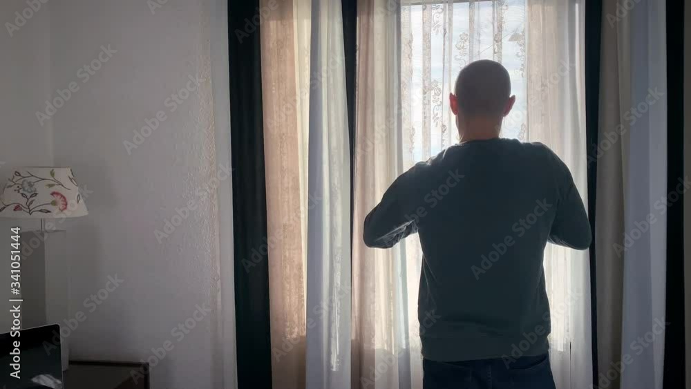 a man stands in a room by the window, opens striped curtains and looks at the city street, the concept of mood, quarantine, morning a man stands in a room by the window, opens striped curtains and looks at the city street, the concept of mood, quarantine, morning