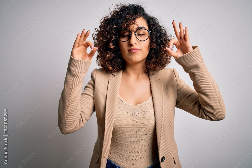 Young curly arab business woman wearing elegant jacket and glasses over white background relax and smiling with eyes closed doing meditation gesture with fingers. Yoga concept. Young curly arab business woman wearing elegant jacket and glasses over white background relax and smiling with eyes closed doing meditation gesture with fingers. Yoga concept.