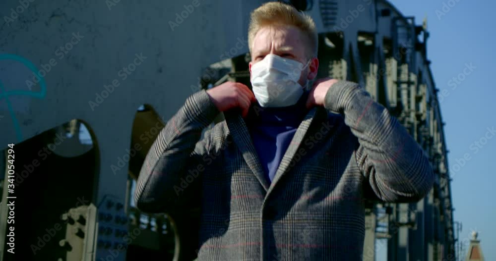 portrait of a man on a city street. he puts a disposable mask on his face, walks along the bridge pillars, frowns and turns up collar portrait of a man on a city street. he puts a disposable mask on his face, walks along the bridge pillars, frowns and turns up collar