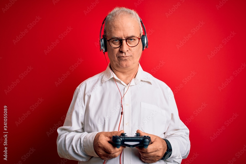 Middle age handsome hoary gamer man playing video game using joystick and headphones with a confident expression on smart face thinking serious Middle age handsome hoary gamer man playing video game using joystick and headphones with a confident expression on smart face thinking serious