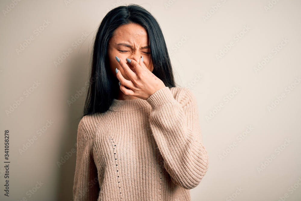 Young beautiful chinese woman wearing casual sweater over isolated white background smelling something stinky and disgusting, intolerable smell, holding breath with fingers on nose. Bad smell Young beautiful chinese woman wearing casual sweater over isolated white background smelling something stinky and disgusting, intolerable smell, holding breath with fingers on nose. Bad smell