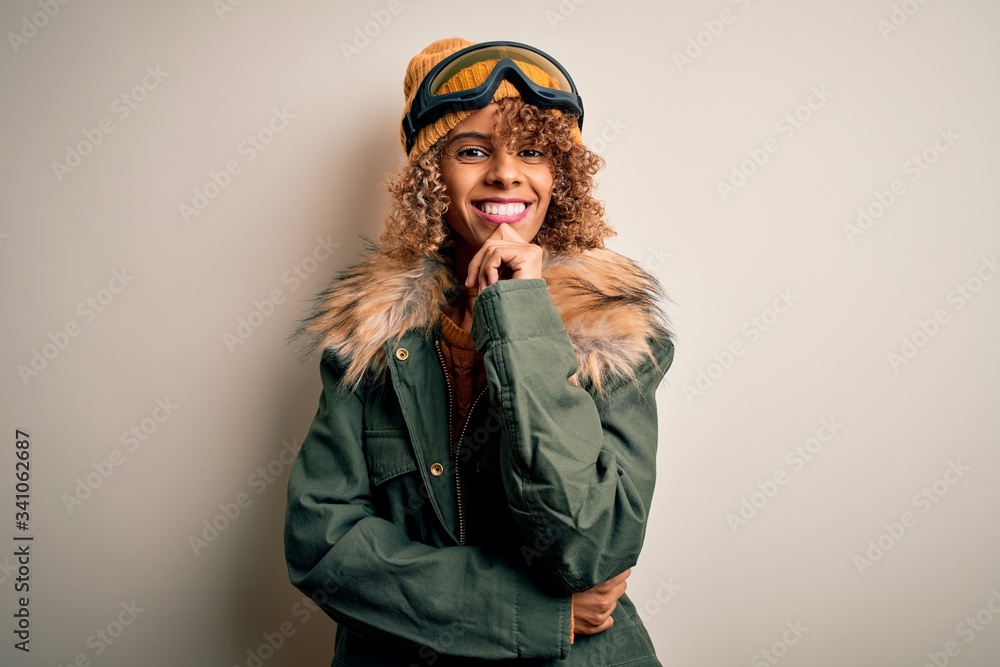 Young african american skier woman with curly hair wearing snow sportswear and ski goggles looking confident at the camera smiling with crossed arms and hand raised on chin. Thinking positive.