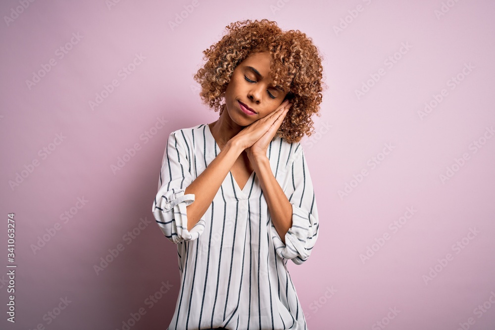 Beautiful african american woman with curly hair wearing striped t-shirt over pink background sleeping tired dreaming and posing with hands together while smiling with closed eyes. Beautiful african american woman with curly hair wearing striped t-shirt over pink background sleeping tired dreaming and posing with hands together while smiling with closed eyes.