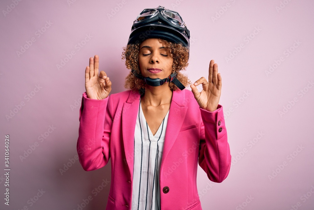 African american motorcyclist woman with curly hair wearing moto helmet over pink background relax and smiling with eyes closed doing meditation gesture with fingers. Yoga concept. African american motorcyclist woman with curly hair wearing moto helmet over pink background relax and smiling with eyes closed doing meditation gesture with fingers. Yoga concept.