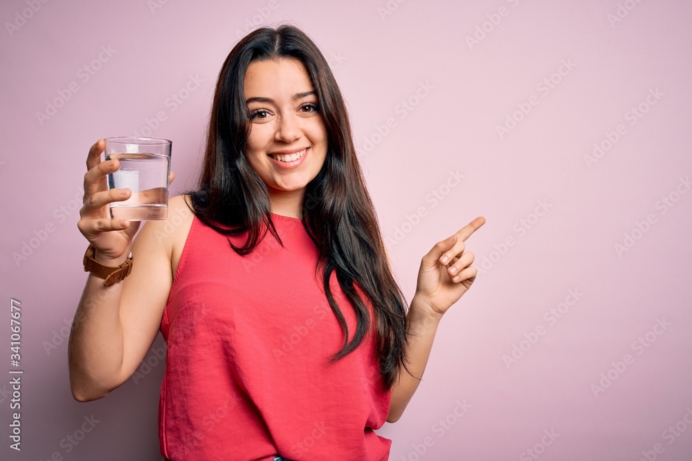 Young brunette woman drinking a glass of fresh water over pink isolated background very happy pointing with hand and finger to the side Young brunette woman drinking a glass of fresh water over pink isolated background very happy pointing with hand and finger to the side