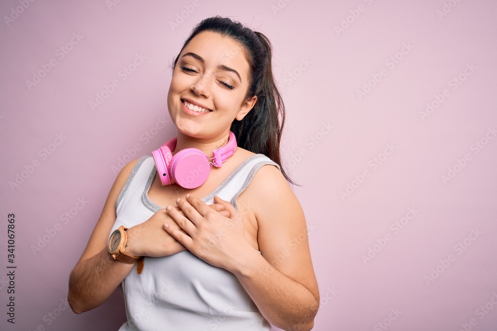 Young brunette fitness woman wearing gym headphones over pink isolated background smiling with hands on chest with closed eyes and grateful gesture on face. Health concept. Young brunette fitness woman wearing gym headphones over pink isolated background smiling with hands on chest with closed eyes and grateful gesture on face. Health concept.